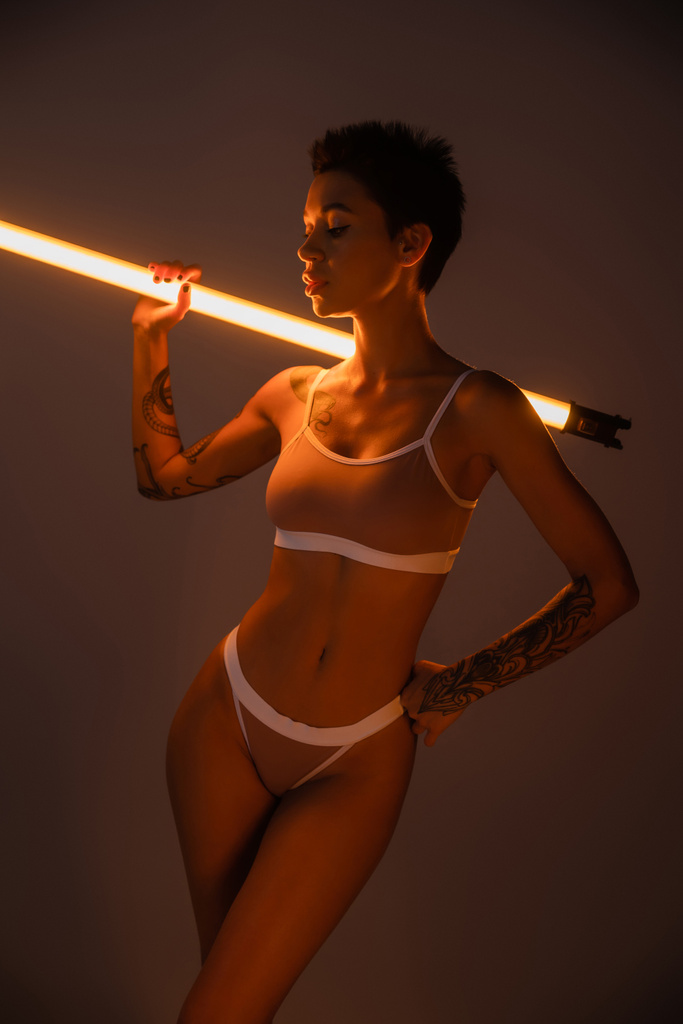 femme tatouée en sous-vêtements tenant lampe fluorescente tout en posant avec la main sur la hanche sur fond sombre - Photo, image