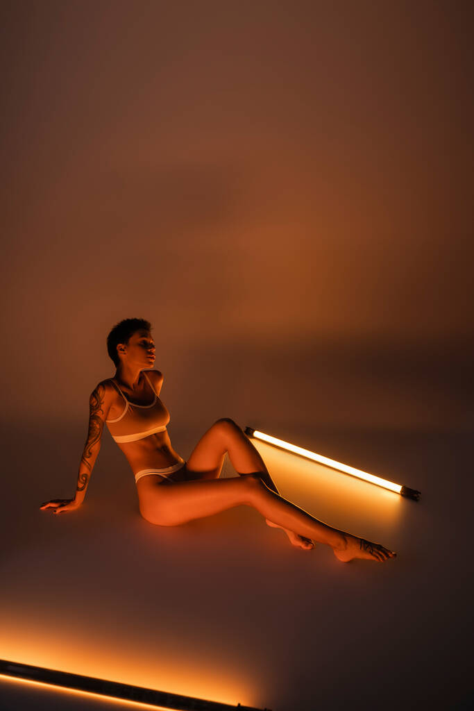 täyspitkä hoikka tatuoitu nainen alusvaatteita istuu lähellä elinvoimainen loistelamput tumma tausta - Valokuva, kuva