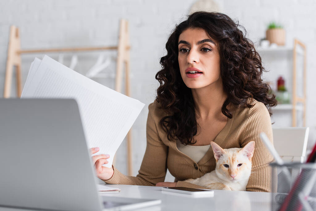 Brunette copywriter holding papers and oriental cat near laptop at home  - Photo, Image