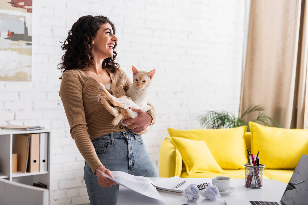 Side view of cheerful copywriter holding oriental cat and papers at home  - Foto, Bild