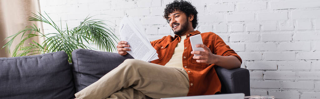 Cheerful indian copywriter looking at papers and holding smartphone on couch at home, banner  - Foto, Imagem