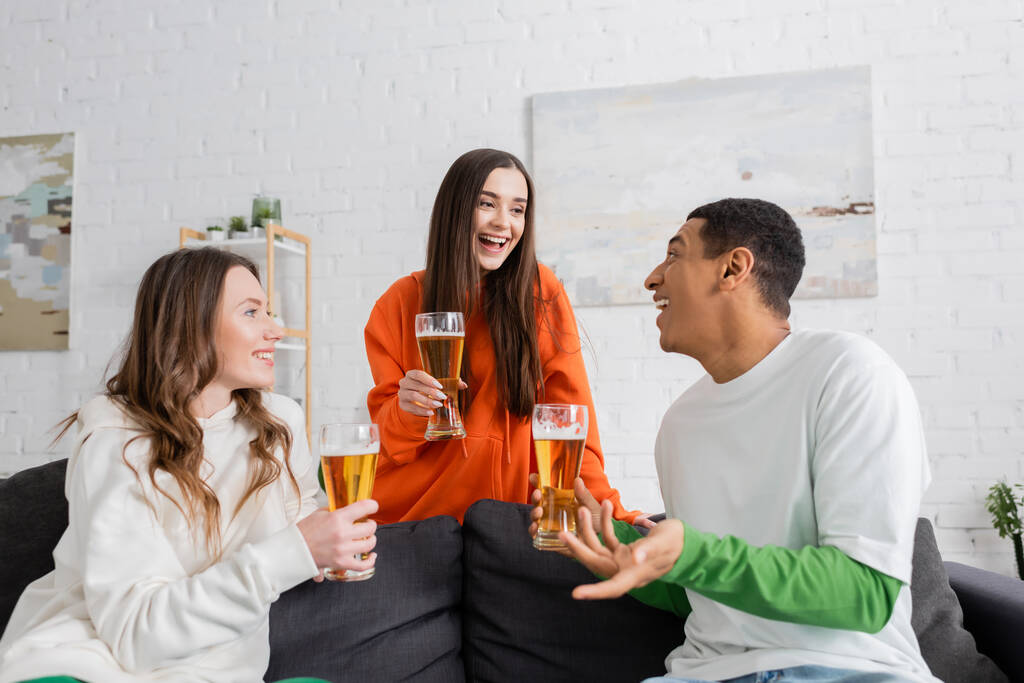 正の女性と幸せなアフリカ系アメリカ人男性がリビングルームでビールを飲みながら  - 写真・画像