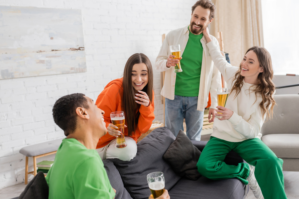 cheerful multiethnic friends holding glasses of beer while chatting in living room  - Photo, Image