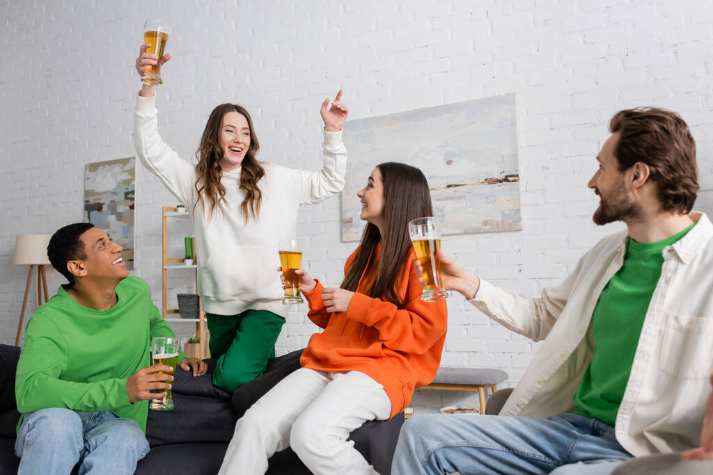 возбужденная женщина со стаканом пива жестикулирует рядом с межрасовыми друзьями в гостиной  - Фото, изображение