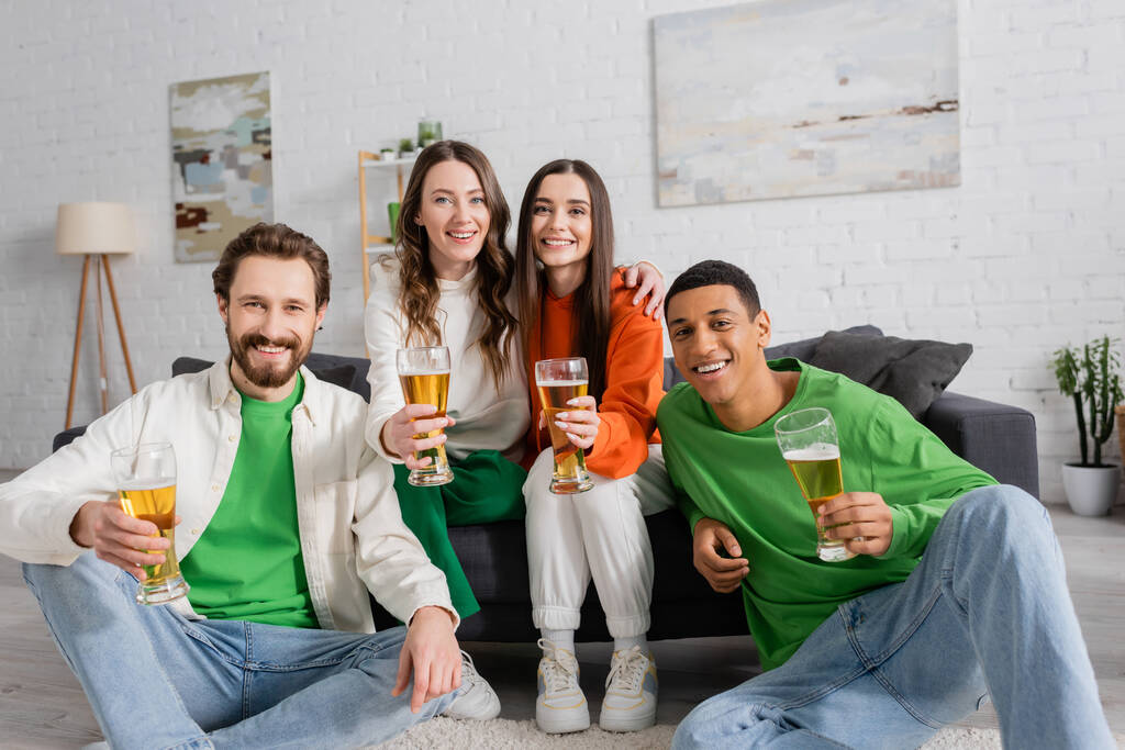 positive and interracial group of friends holding glasses of beer in living room  - Foto, Imagem