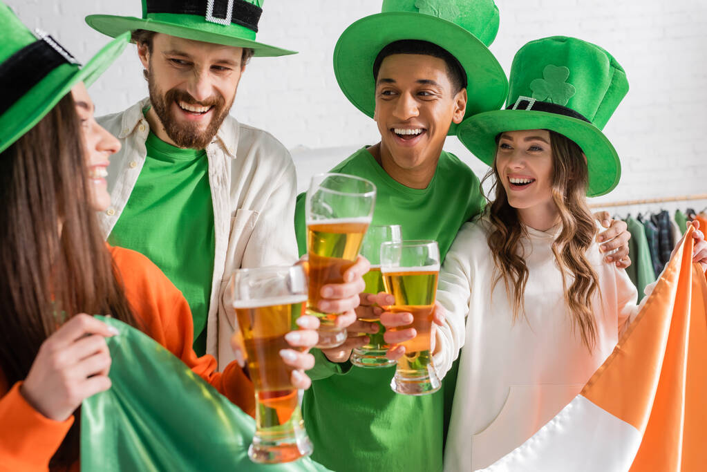 счастливые и межрасовые друзья в зеленых шляпах, держащие бокалы пива и ирландский флаг во время празднования Дня Святого Патрика  - Фото, изображение