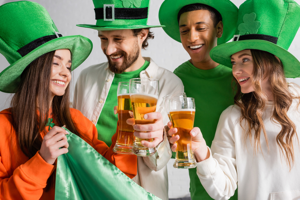 cheerful and interracial friends in green hats clinking glasses of beer near Irish flag while celebrating Saint Patrick Day  - Foto, imagen