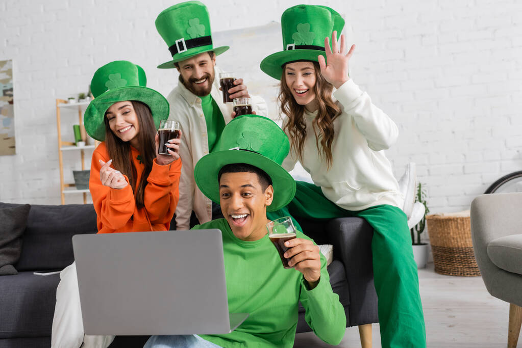 joyful african american man holding laptop during video call near cheerful friends with glasses of beer on Saint Patrick Day - Photo, Image