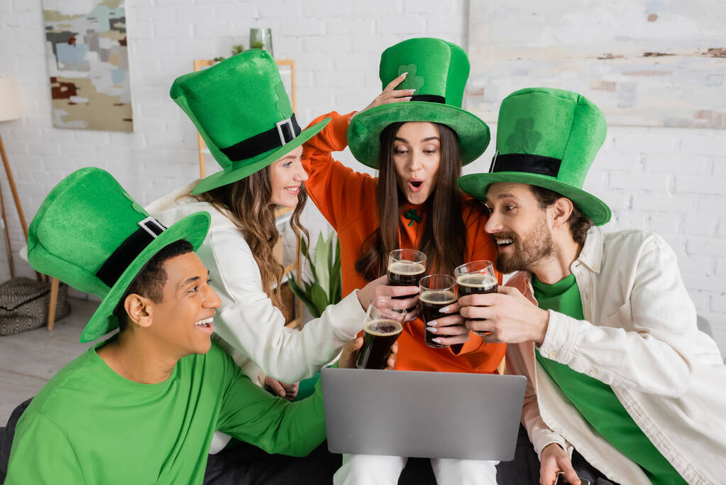 excited woman looking at laptop during video call while toasting glasses of beer with interracial friends on Saint Patrick Day - Valokuva, kuva