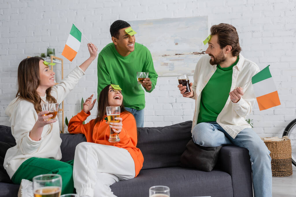 gelukkig interraciale vrienden met plakkerige notities op voorhoofden houden drankjes en Ierse vlaggen tijdens het spelen Raad eens wie spel op Saint Patrick Day - Foto, afbeelding