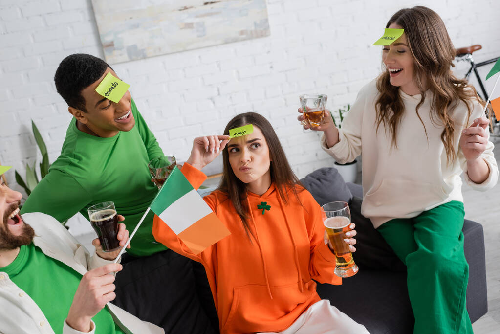 multiethnische Freunde mit klebrigen Zetteln auf der Stirn, die Alkoholgetränke in der Hand halten und nachdenkliche Frauen beim Ratespiel anschauen  - Foto, Bild