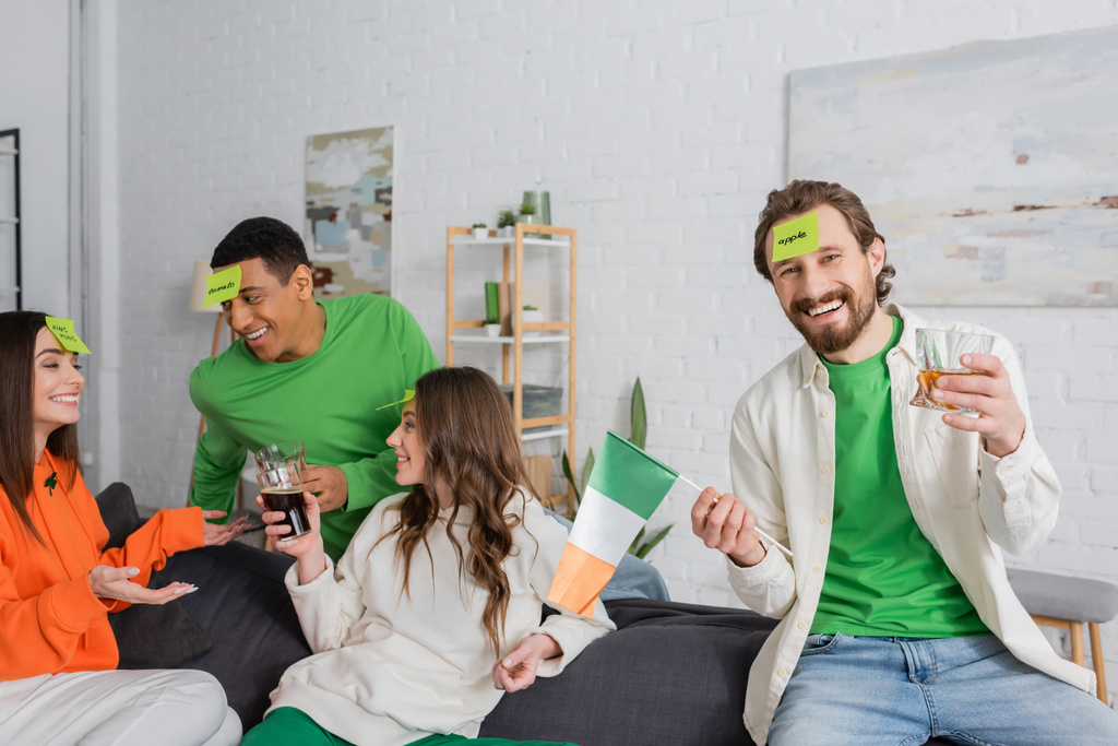 happy bearded man with apple word on sticky note holding Irish flag and drink near interracial friends on Saint Patrick Day - Foto, imagen