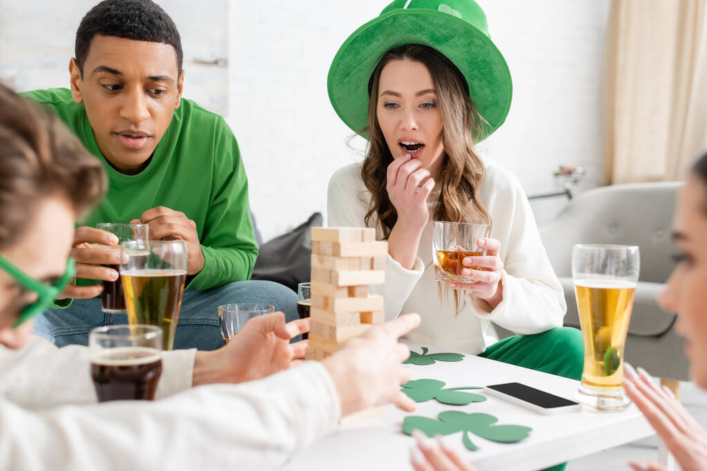 Pensativo interracial amigos con cerveza jugando bloques de madera juego mientras se celebra santo patrick día en casa  - Foto, imagen