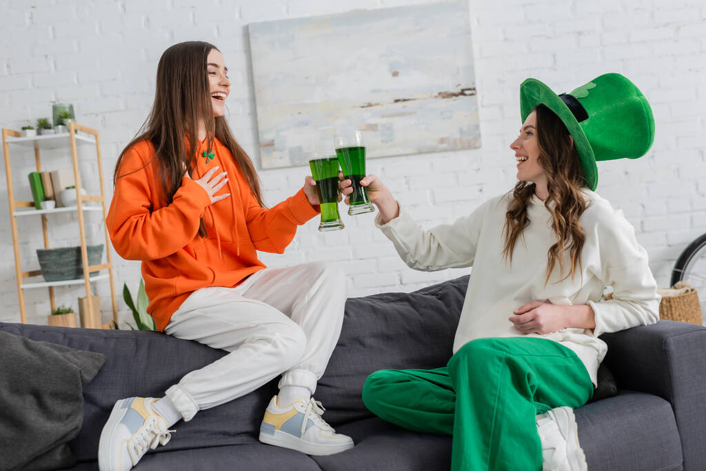 Cheerful young women clinking green beer and talking on couch on saint patrick day  - Foto, afbeelding