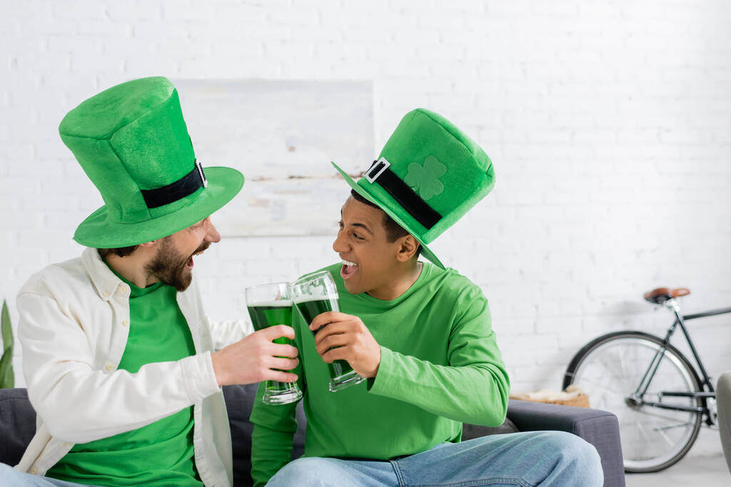 Excited multiethnic friends in green hats clinking beer during saint patrick day at home  - Photo, image