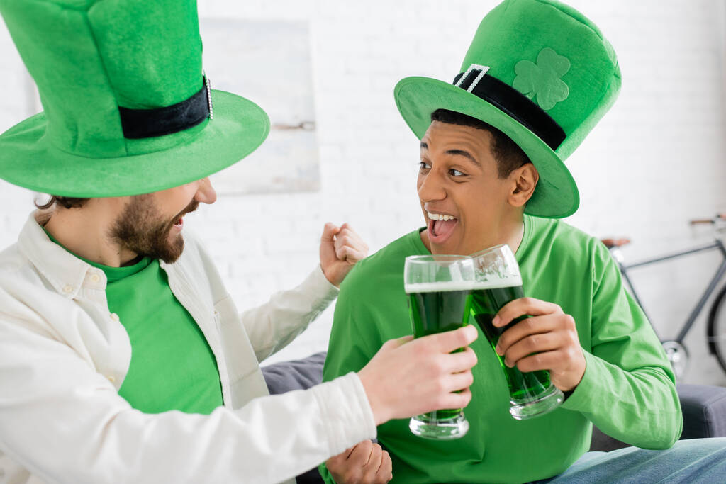 Excited interracial friends clinking green beer during saint patrick day at home  - Photo, image