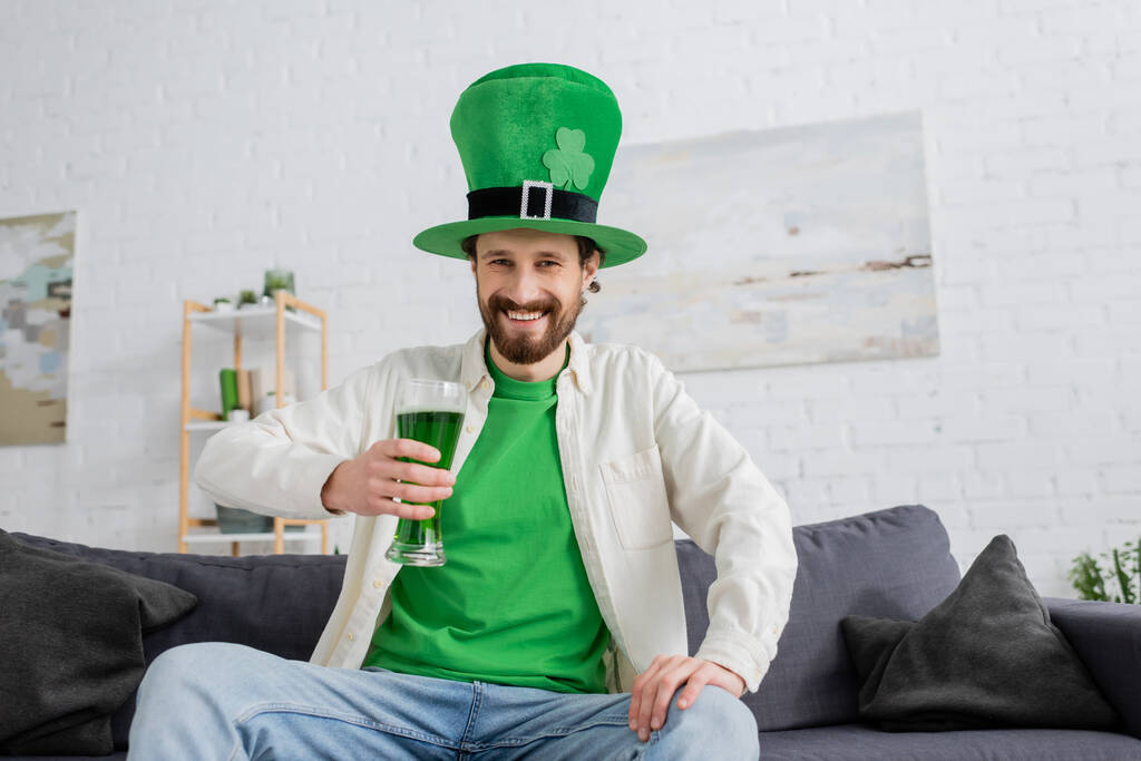 Hymyilevä mies vihreässä hatussa pitäen olutta ja katsoen kameraa pyhimys patrickin juhlan aikana kotona  - Valokuva, kuva
