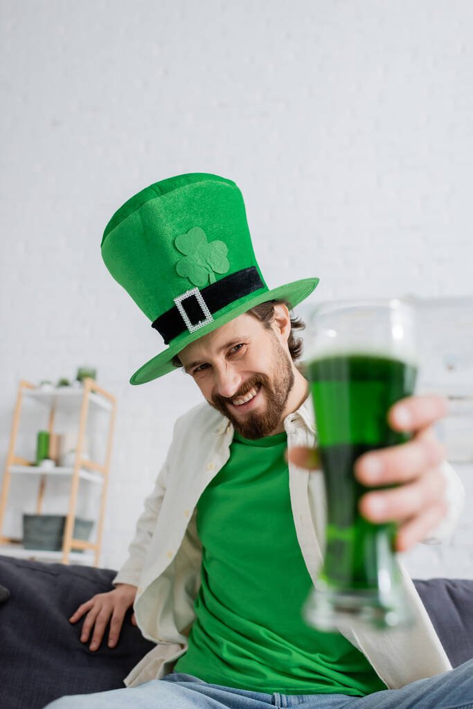 Smiling man in hat holding blurred green beer during saint patrick celebration at home  - Foto, imagen