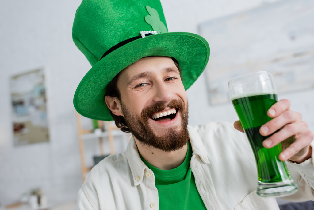 Cheerful man in saint patrick hat holding green beer and looking at camera at home  - Photo, Image