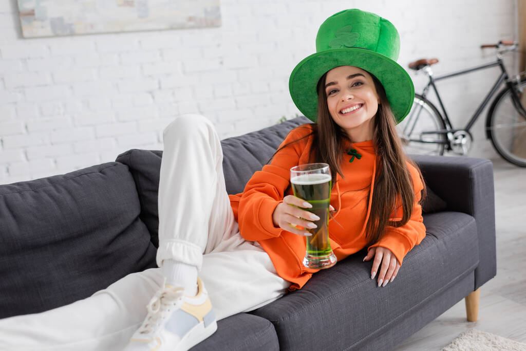 Sorrindo jovem mulher em chapéu verde segurando cerveja enquanto deitado no sofá  - Foto, Imagem