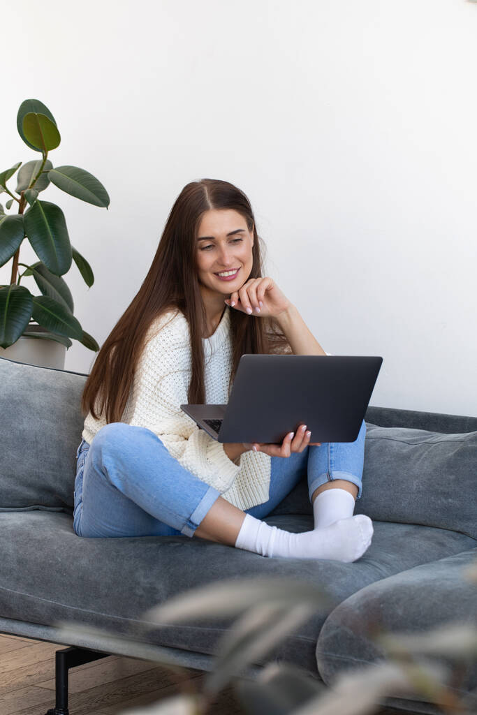 Młoda kobieta patrzy na monitor laptopa i uśmiecha się, siedząc na kanapie w salonie otoczona roślinami domowymi. - Zdjęcie, obraz