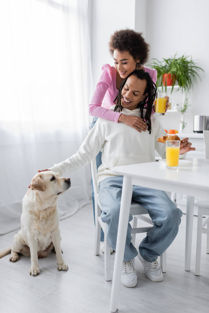 Mosolygó afro-amerikai nő ölelés barátja közelében labrador és reggeli a konyhában  - Fotó, kép