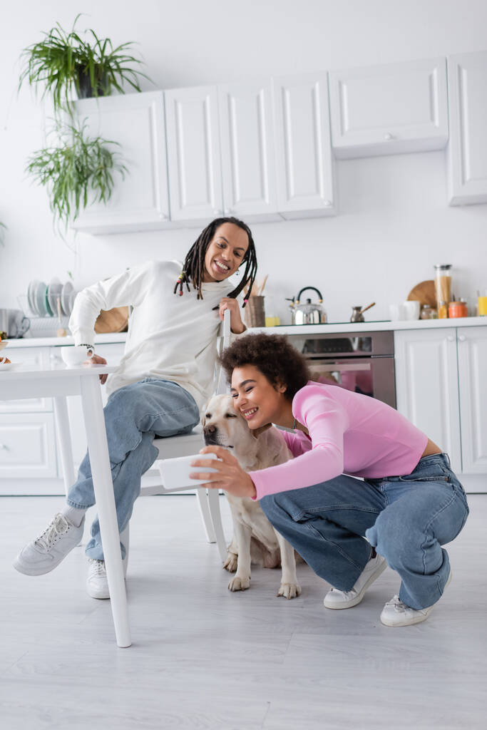 Sourire femme afro-américaine prenant selfie avec labrador près de petit ami dans la cuisine  - Photo, image