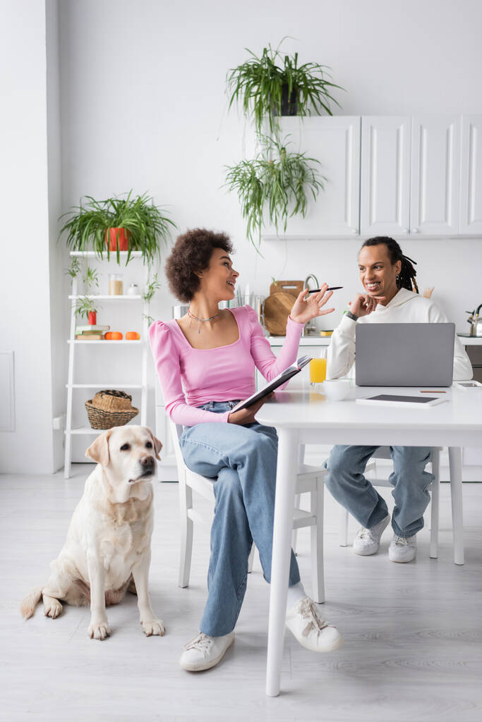 Smiling african american woman with notebook talking to boyfriend near devices and labrador at home  - Photo, Image