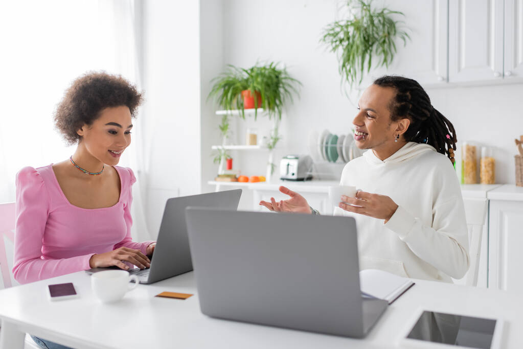 Femme afro-américaine positive utilisant un ordinateur portable pendant que son petit ami parle à la maison  - Photo, image