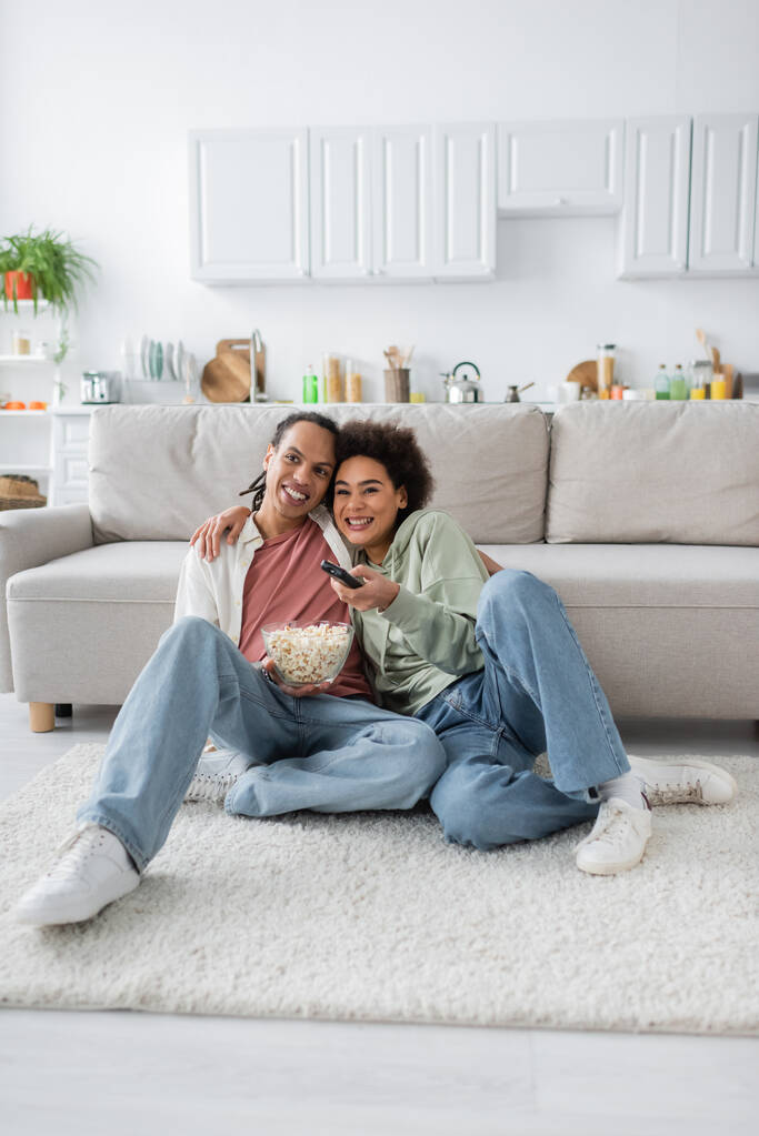 Smiling african american woman hugging boyfriend with popcorn while watching tv at home  - Zdjęcie, obraz