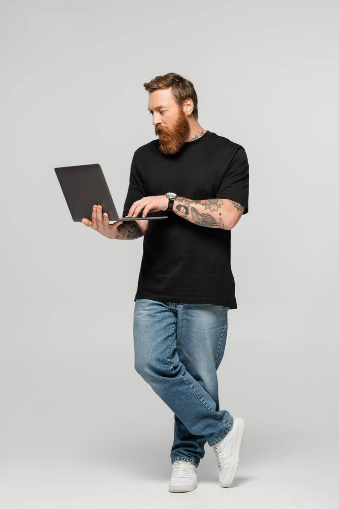 повна довжина зосередженого чоловіка в джинсах і чорній футболці з ноутбуком на сірому фоні
 - Фото, зображення