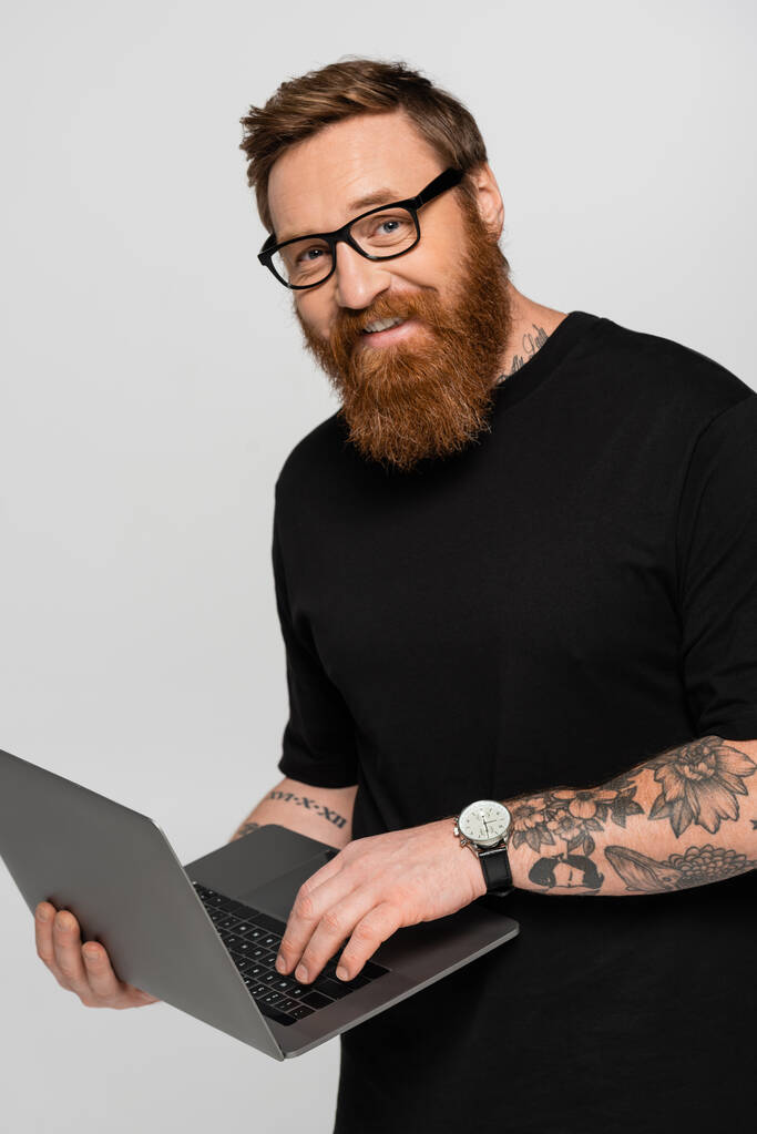 bearded freelancer in eyeglasses holding laptop and smiling at camera isolated on grey - Zdjęcie, obraz