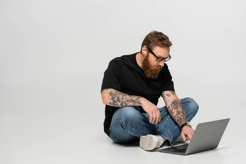 skoncentrowany brodaty mężczyzna w okularach za pomocą laptopa podczas siedzenia ze skrzyżowanymi nogami na szarym tle - Zdjęcie, obraz