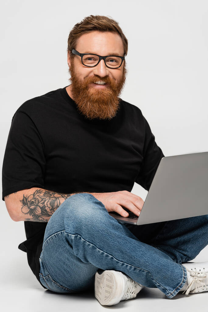 happy bearded freelancer holding laptop while sitting with crossed legs on grey background - Zdjęcie, obraz