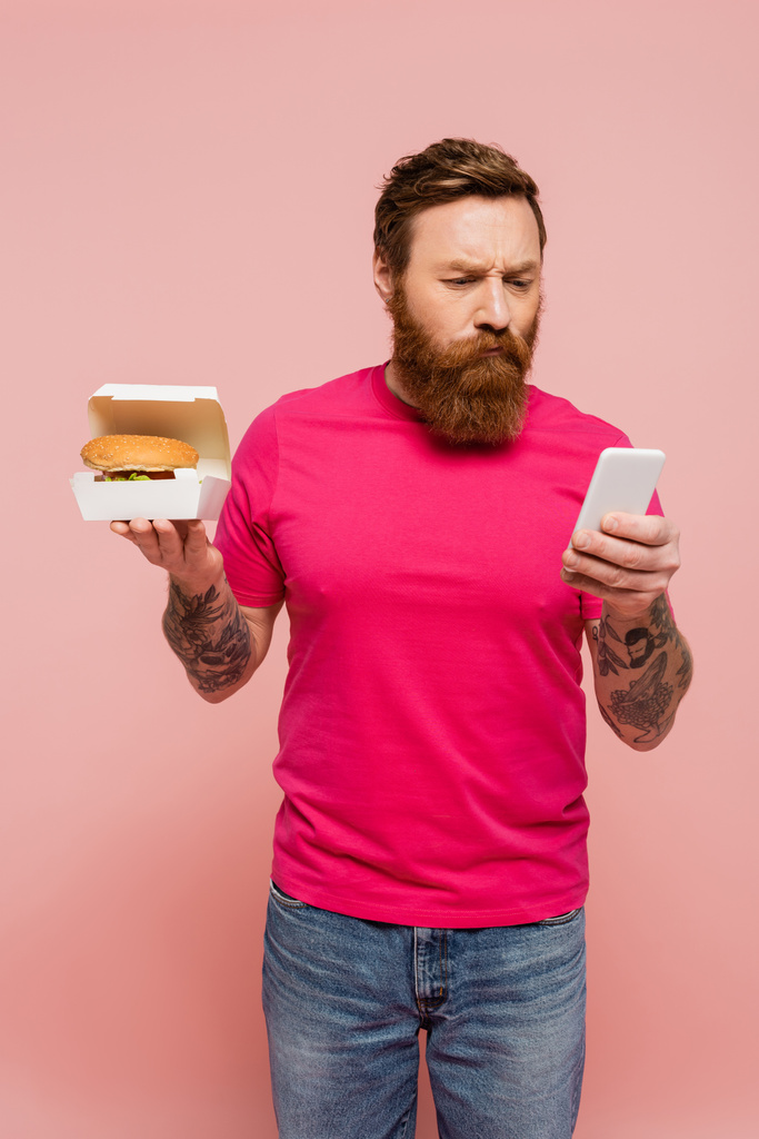 thoughtful bearded man holding carton box with hamburger and using mobile phone isolated on pink - Foto, Imagem
