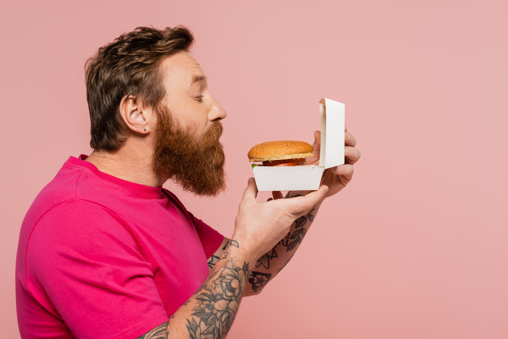 боковой вид на бородатого татуированного человека, нюхающего вкусный бургер в коробке упаковки изолированы на розовый - Фото, изображение