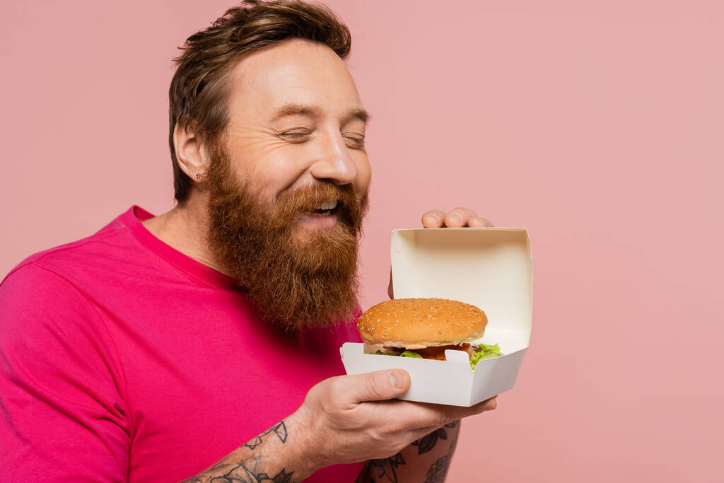 hombre barbudo complacido con los ojos cerrados sosteniendo paquete de cartón con sabrosa hamburguesa aislada en rosa - Foto, Imagen