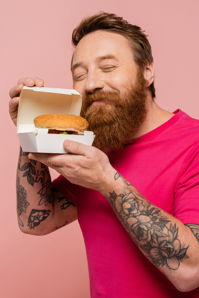 pleased stylish man with closed eyes smelling tasty burger in carton pack isolated on pink - 写真・画像