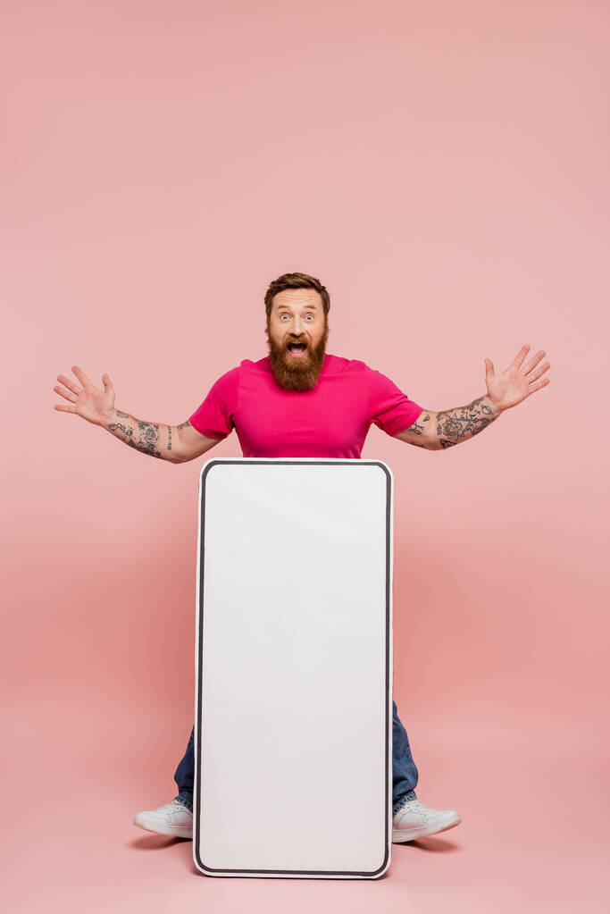 izgatott tetovált férfi bemutató wow gesztus közelében hatalmas mobiltelefon sablon rózsaszín háttér - Fotó, kép