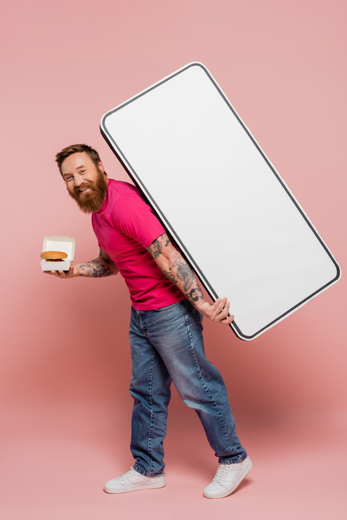 happy bearded man in magenta t-shirt and jeans carrying hamburger and white template of smartphone on pink background - 写真・画像