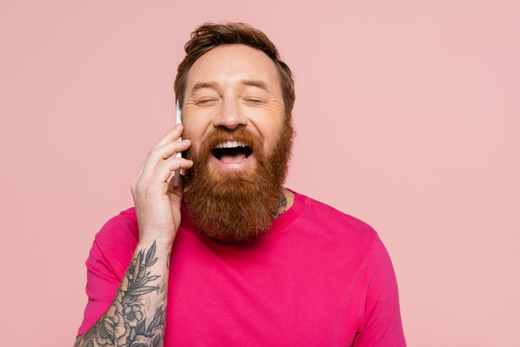 喜びに満ちた髭の男が携帯電話で目を閉じて笑ってピンクに隔離された - 写真・画像