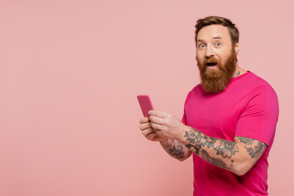 hombre barbudo excitado con la boca abierta usando el teléfono celular y mirando la cámara aislada en rosa - Foto, imagen