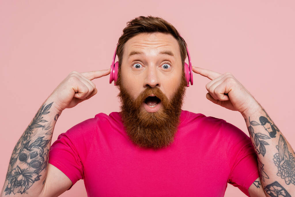 изумленный бородатый мужчина в пурпурной футболке указывает на беспроводные наушники и смотрит на камеру, изолированную на розовом - Фото, изображение