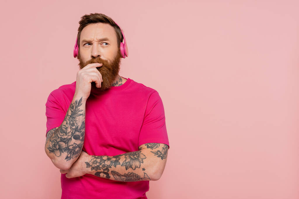 στοχαστικός άνθρωπος με τατουάζ σε ασύρματα ακουστικά κρατώντας το χέρι κοντά στο πρόσωπο και κοιτάζοντας μακριά απομονώνονται σε ροζ - Φωτογραφία, εικόνα