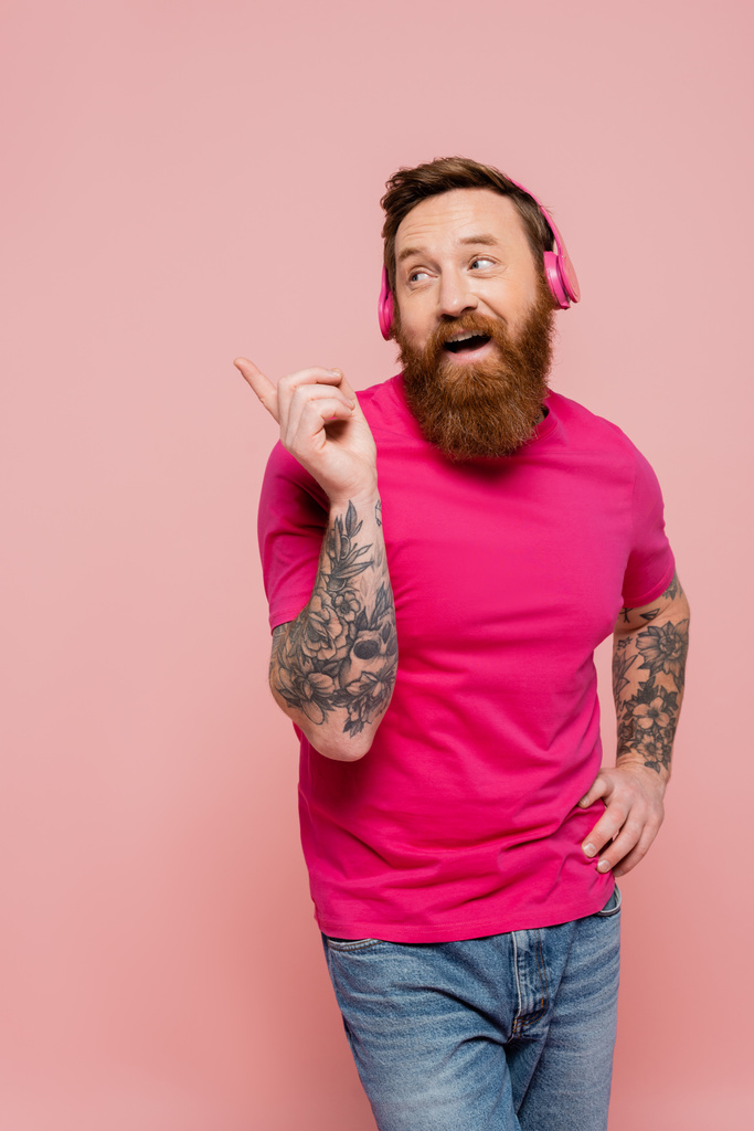 Pembe tişörtlü ve kablosuz kulaklıklı neşeli sakallı adam pembe üzerine izole edilmiş parmağıyla işaret ediyor. - Fotoğraf, Görsel
