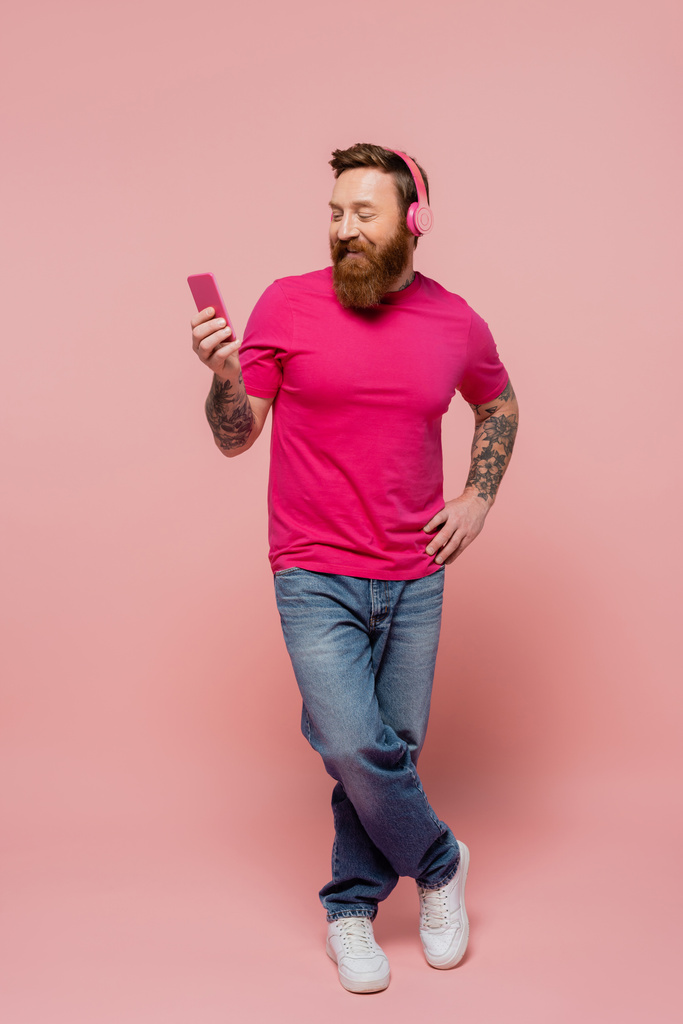 longitud completa de hombre feliz en auriculares inalámbricos que sostienen el teléfono inteligente mientras está de pie con la mano en la cadera sobre fondo rosa - Foto, imagen
