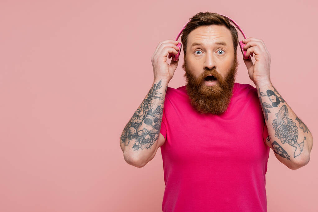 surprised tattooed man with open mouth holding wireless headphones while looking at camera isolated on pink - Fotografie, Obrázek