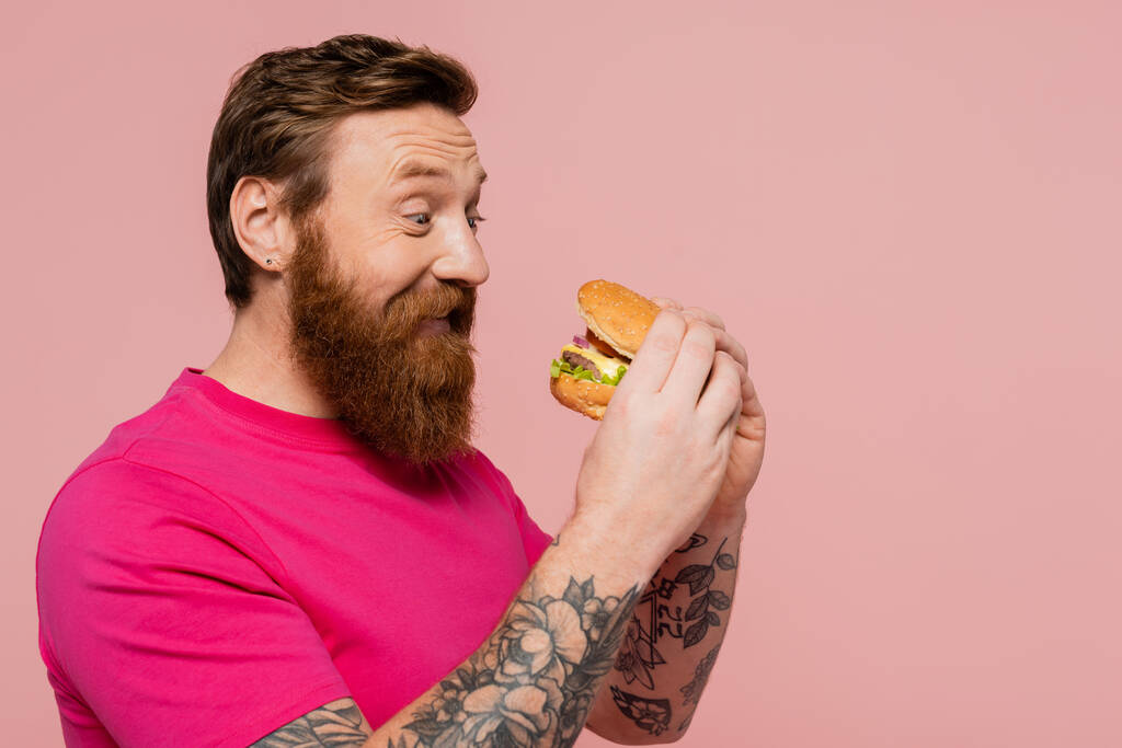 ピンク色に隔離されたおいしいハンバーガーを持ったマゼンタTシャツの幸せな髭男 - 写真・画像