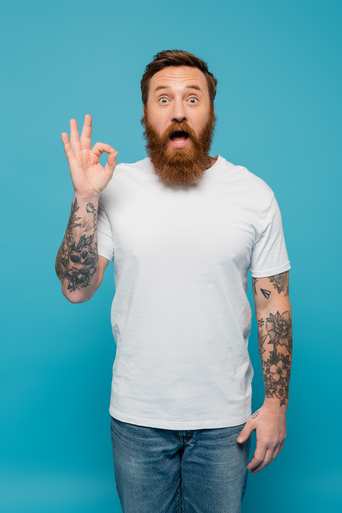 впечатленный бородатый мужчина в белой футболке смотрит в камеру и показывает хорошо знак изолирован на синий - Фото, изображение