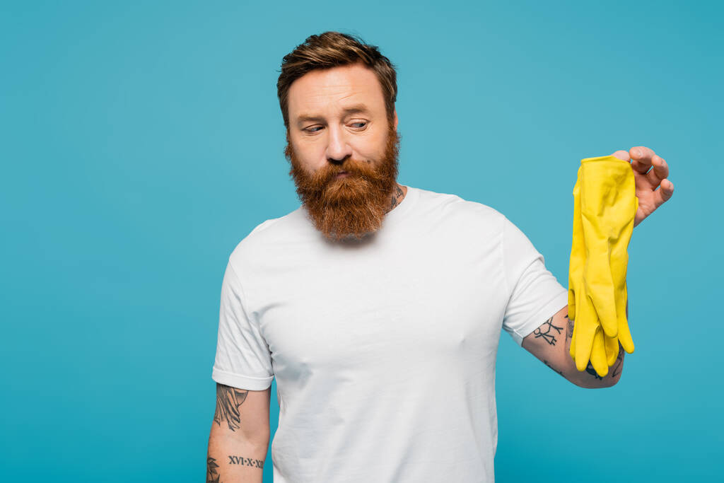 displeased bearded man in white t-shirt holding yellow rubber gloves isolated on blue - Foto, Bild